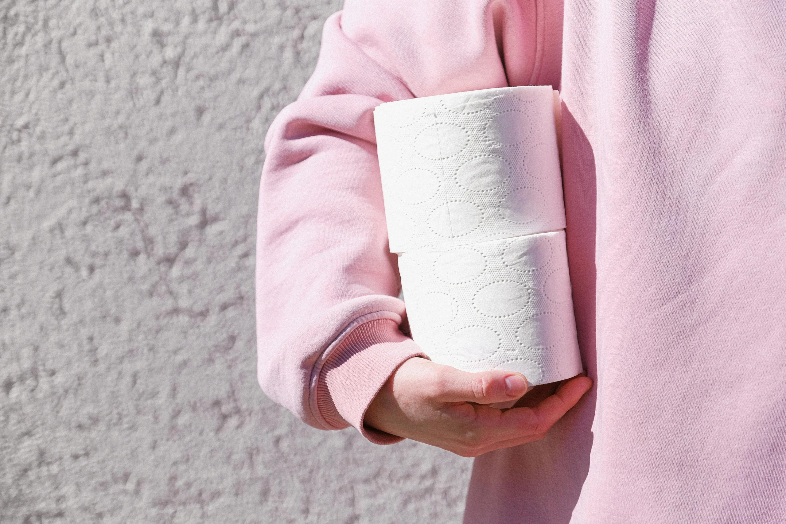 Close-up of a person in a pink sweater holding two toilet paper rolls outdoors.