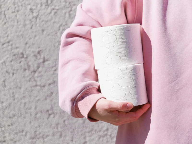 Close-up of a person in a pink sweater holding two toilet paper rolls outdoors.