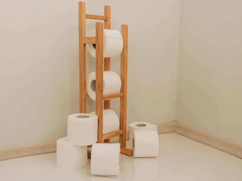 Neatly stacked toilet paper rolls on a bamboo rack in a minimalist bathroom corner.