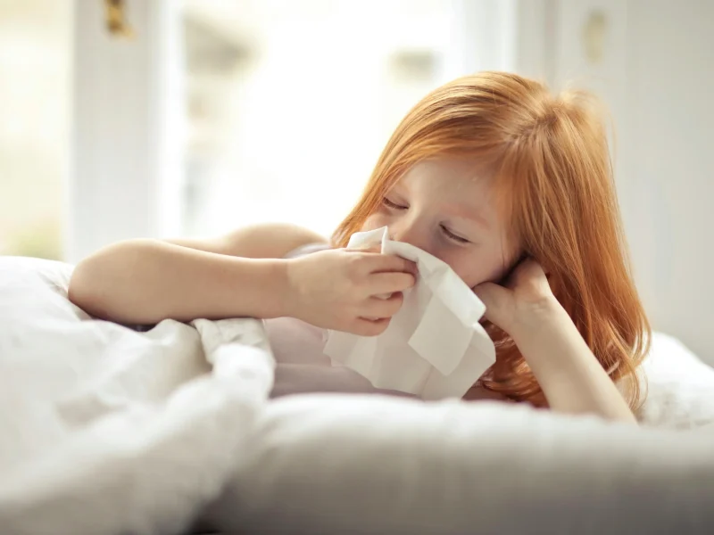 Small sick redhead kid with eyes closed cleaning nose with white tissue paper while lying alone in bed in light modern bedroom in daytime
