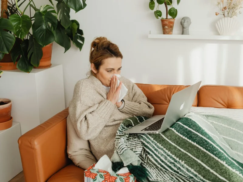 Woman in cozy sweater using laptop on sofa, surrounded by indoor plants, while blowing nose.
