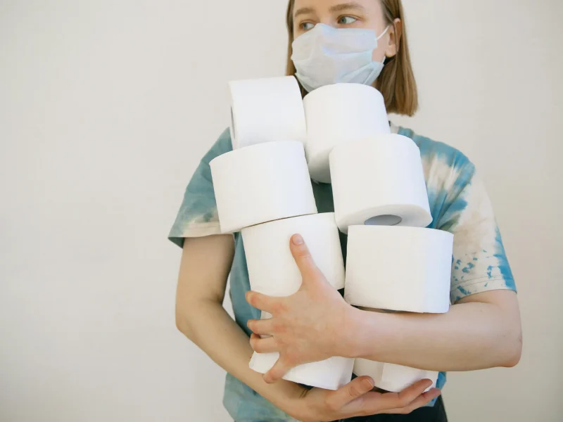 Woman wearing a face mask holding several toilet paper rolls indoors.